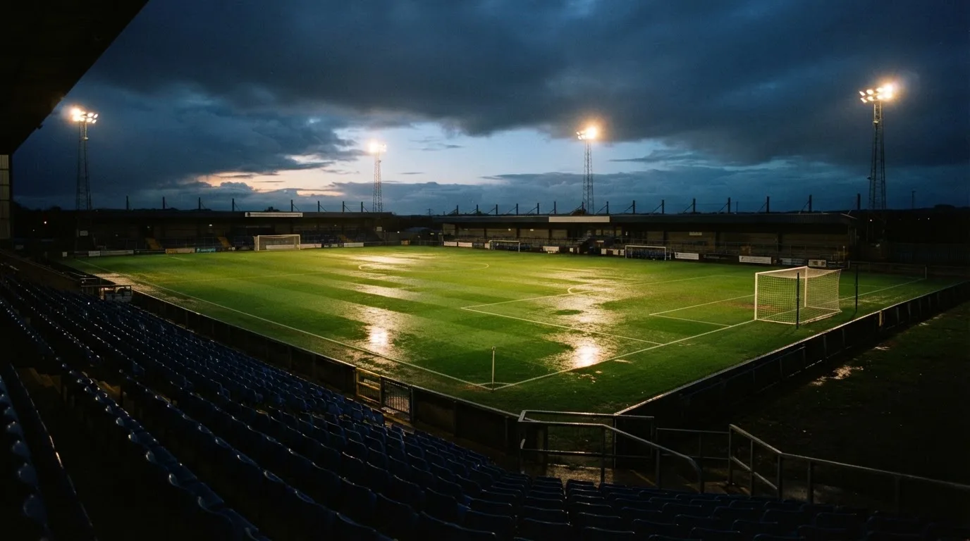 Ein Fußballplatz mit Flutlichtmasten in der Abenddämmerung, aus der Ferne gesehen, leere Tribünen und nasser Rasen unter bewölktem Himmel.