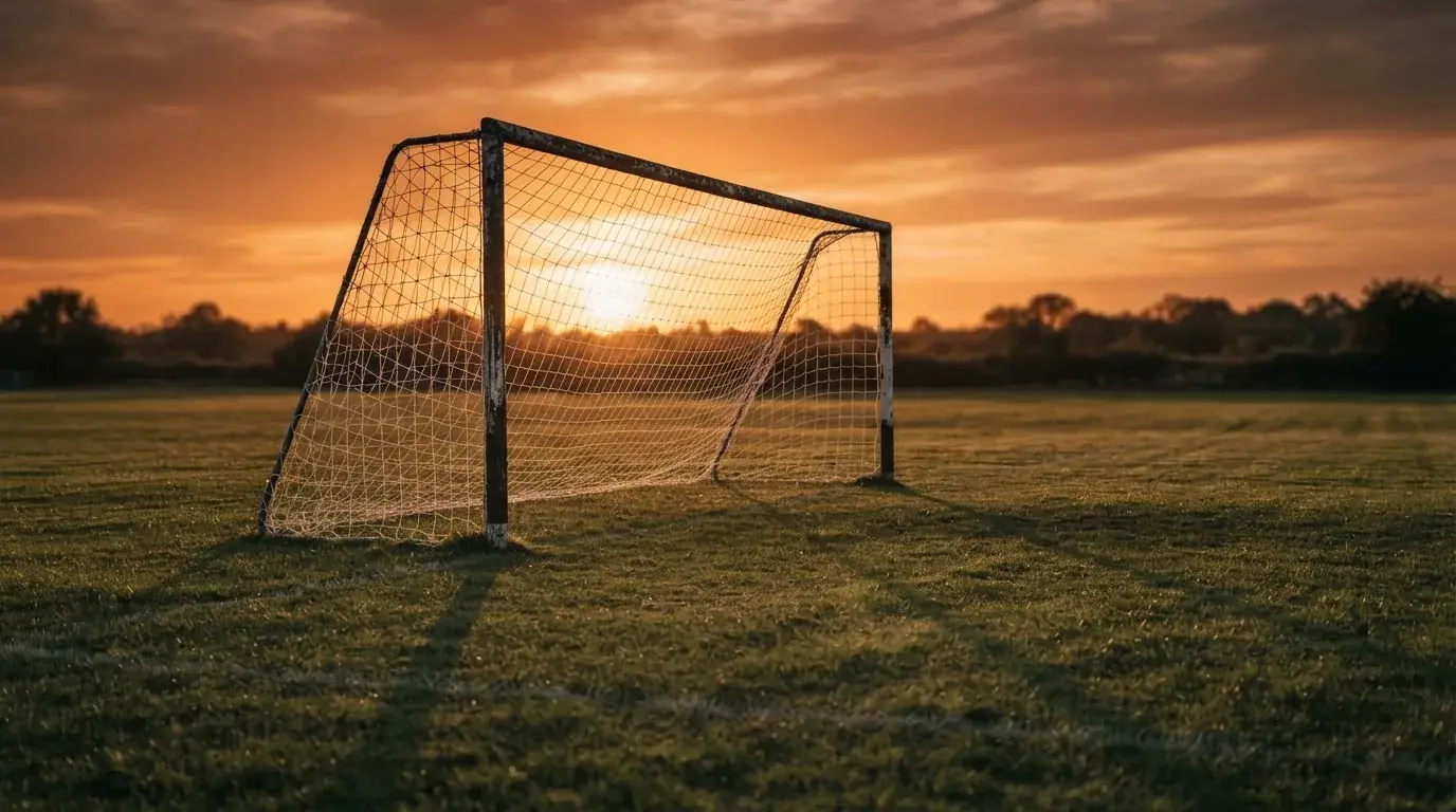 Ein Fußballtor mit Netz bei Sonnenuntergang auf einem leeren Platz, die Schatten der Torpfosten liegen lang auf dem Gras.