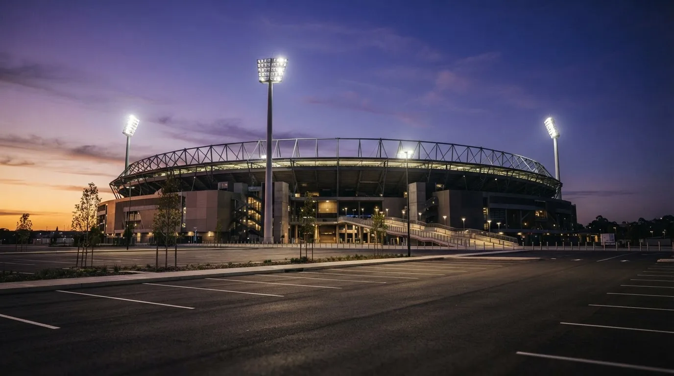 Ein Fußballstadion von außen in der Dämmerung, hohe Flutlichtmasten vor einem violetten Himmel, davor ein leerer Parkplatz.