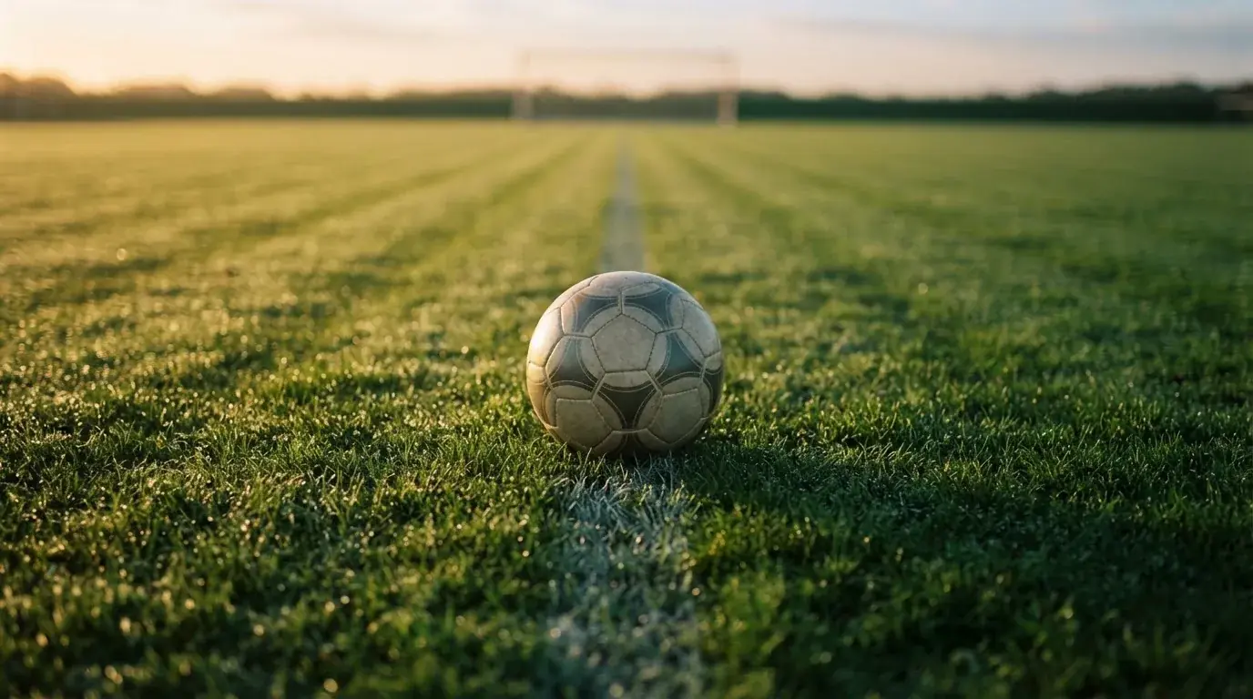 Ein Fußball liegt auf dem Anstoßpunkt eines Rasens bei Sonnenaufgang, Tau glitzert auf den Halmen.