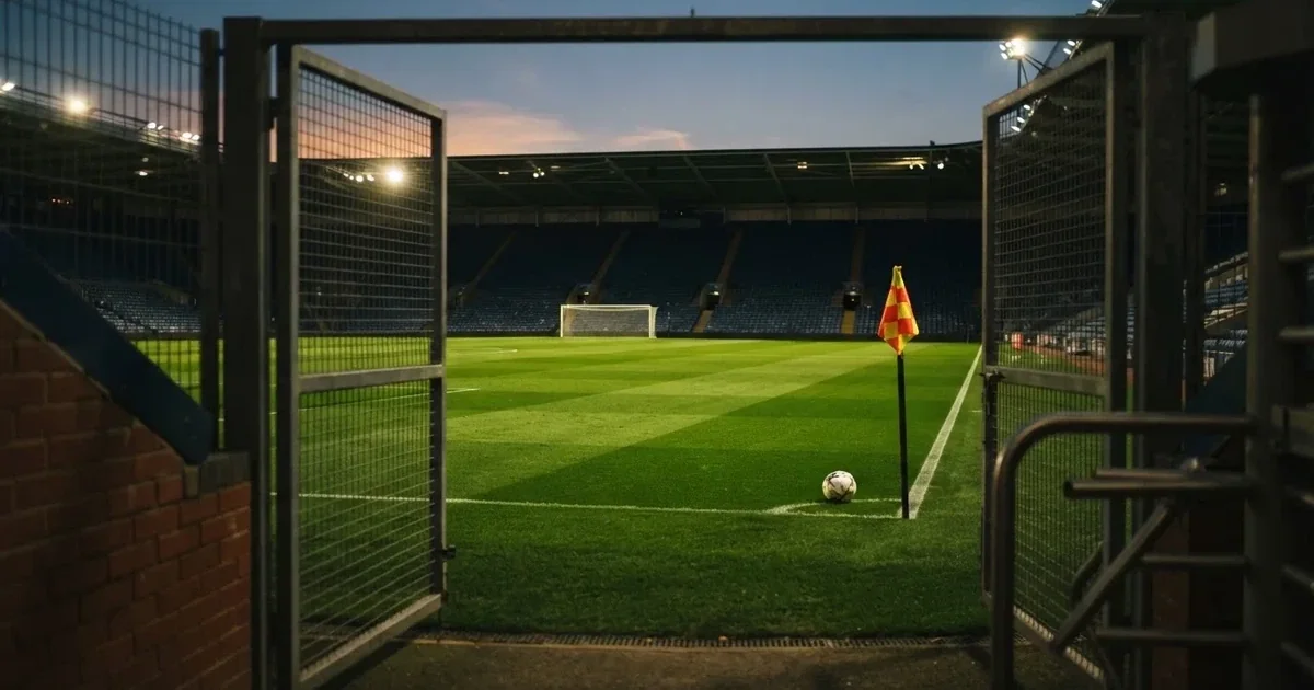 Blick durch ein Stadiontor auf einen grünen Rasen im Abendlicht, ein Ball ruht an der Eckfahne, die Tribünen sind leer.