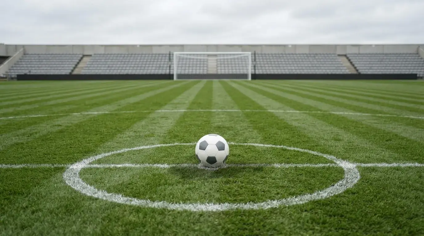 Ein Fußball liegt auf dem Anstoßpunkt eines leeren Rasenplatzes, im Hintergrund eine ruhige Tribüne unter bedecktem Himmel.
