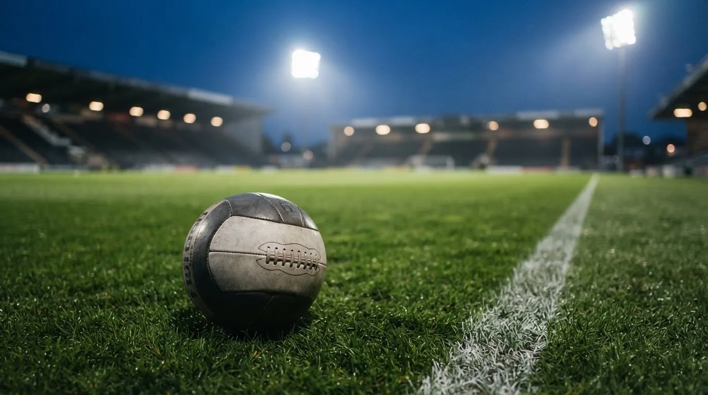 Ein Fußball liegt auf dem Rasen eines leeren Platzes, im Hintergrund ein Stadion unter Flutlicht in der Abenddämmerung.