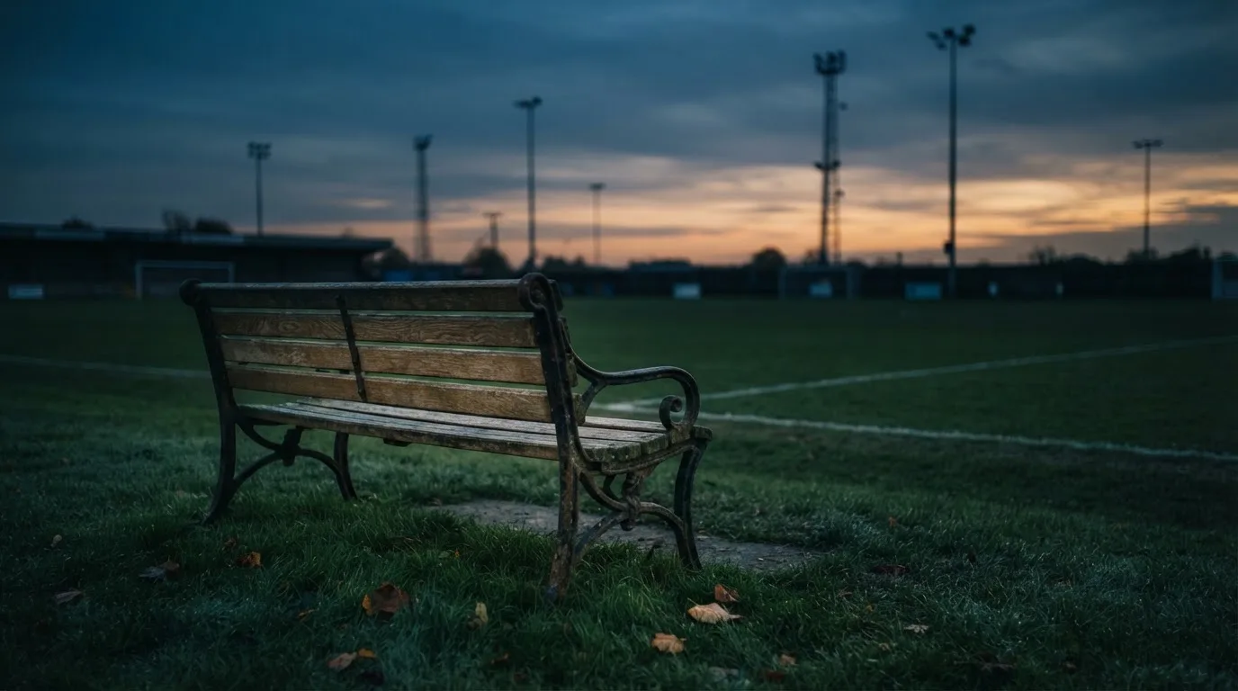 Eine leere Parkbank am Rand eines Fußballfelds in der Abenddämmerung, im Hintergrund ragen dunkle Flutlichtmasten in den Himmel.