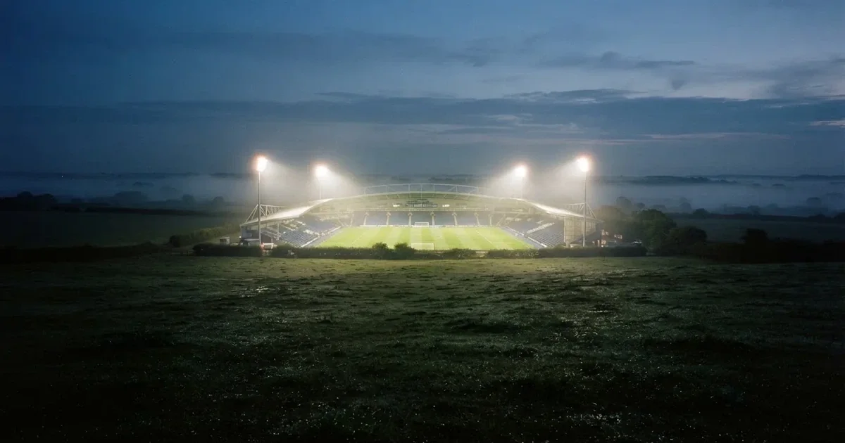 Ein Stadion unter Flutlicht, aus großer Entfernung über eine dunkle Wiese fotografiert, am Himmel zieht Nebel auf.