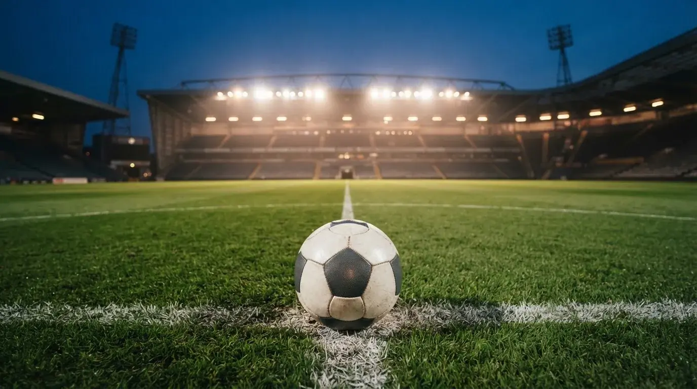 Ein Fußball liegt auf dem Anstoßpunkt eines leeren Rasenplatzes, im Hintergrund steht ein Stadion unter Flutlicht in der Dämmerung.
